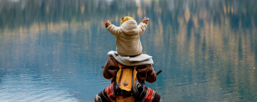 Fotografie einer Person, die ein Kind auf den Schultern trägt und auf einen See blickt, in dem sich die umstehenden Bäume spiegeln.
