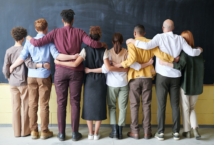 Rückenansicht von 8 farbenfroh gekleideten Personen, die vor einer Tafel stehen und freundschaftlich ihre Arme umeinander legen.