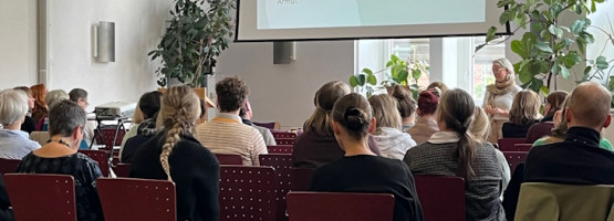 Fotografie von hinten, auf der Seminarteilnehmende zu sehen sind, die auf eine Leinwand blicken, auf der Seminarinhalte der Fachtagung Familienbildung 2023 präsentiert werden.