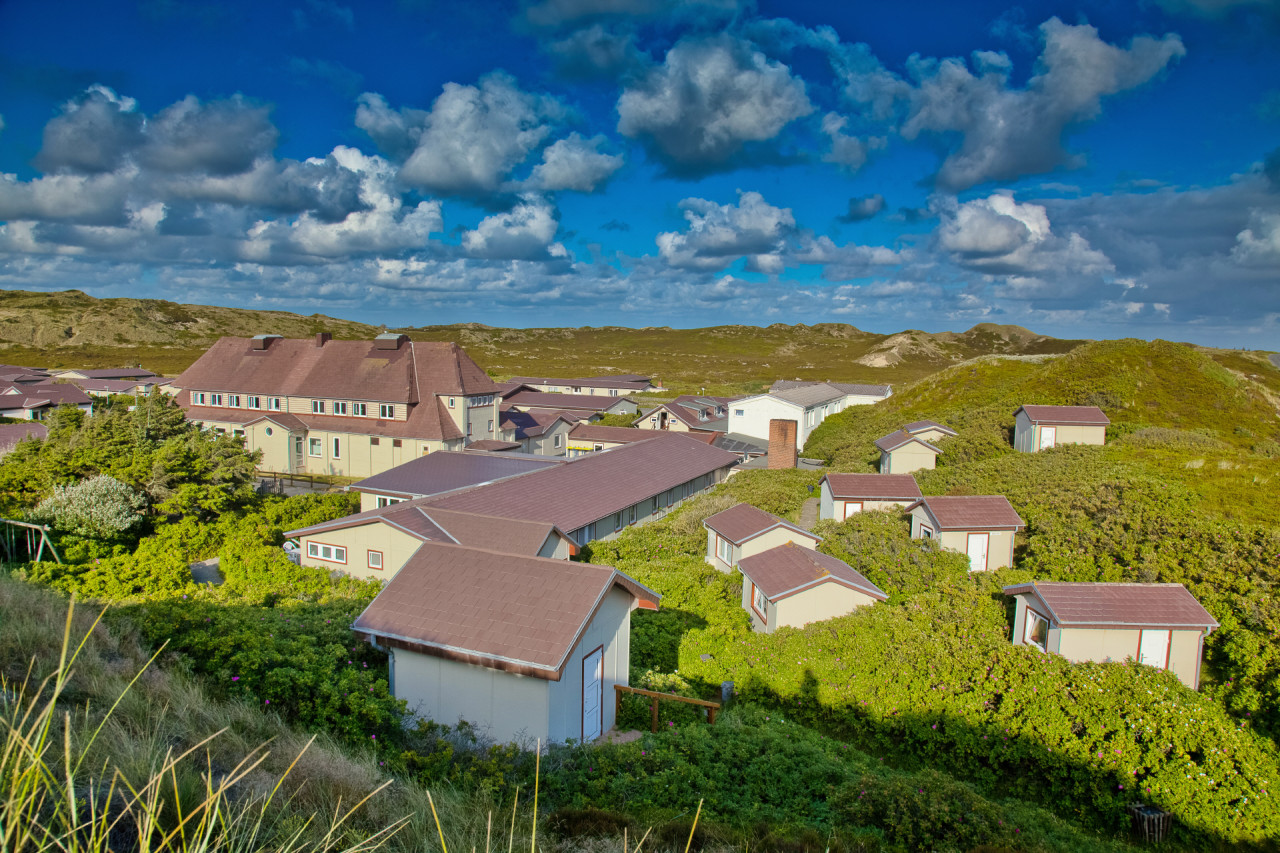 Blick auf das Klappholttal, man sieht ein grünes Tal im Sonnenschein voller Büsche, in welchen kleine Ferienhäuser liegen und ein großes Seminarhaus. Der Himmel ist blau mit kleinen Wölkchen.