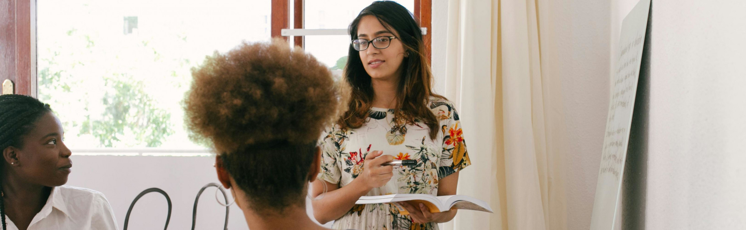 Eine Person steht mit einem aufgeklappten Buch und Stift in der Hand vor einem Plakat an der Wand und erklärt zwei weiteren Personen etwas.