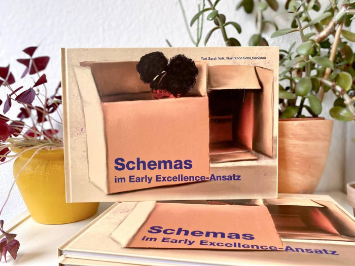 Das Schemabuch lehnt an zwei Zimmerpflanzen. Unter dem stehenden Buch, liegt ein weiteres Buch.